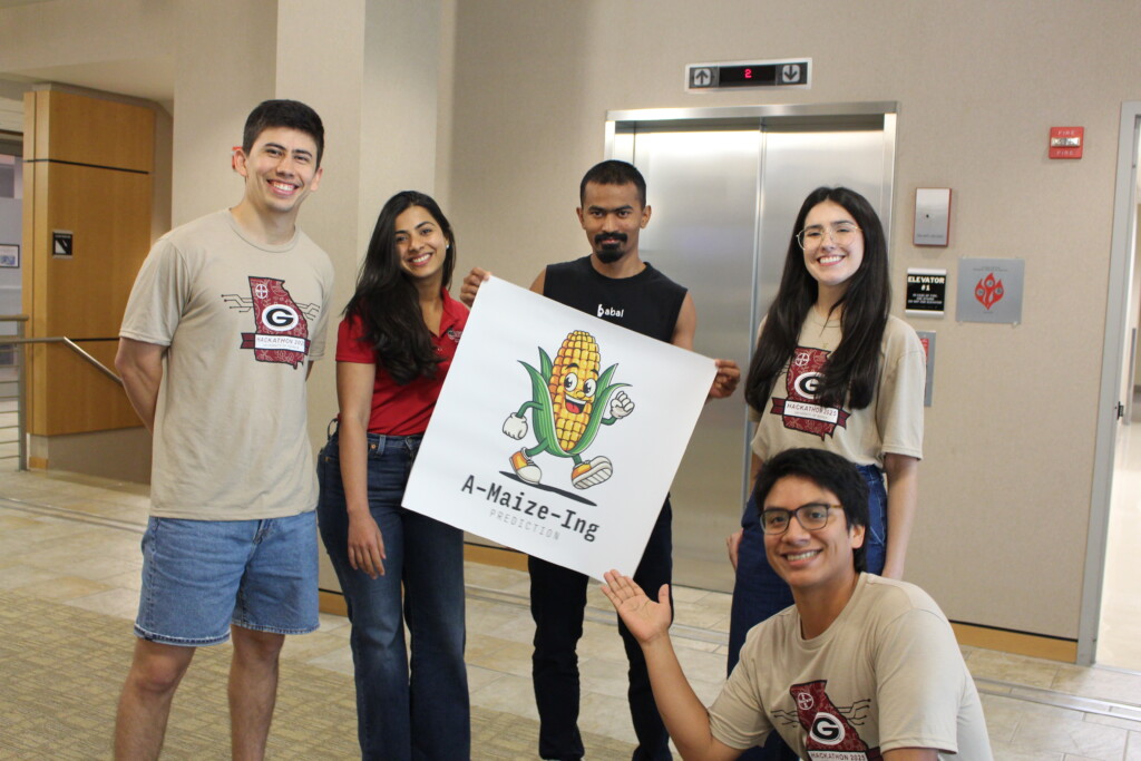 four students stand in a row in a tan hallway in front of an elevator and hold a square poster with a cartoon corn and a logo that says "A-Maize-Ing Prediction" at the bottom. One student kneels in front of the other student and points toward the poster.