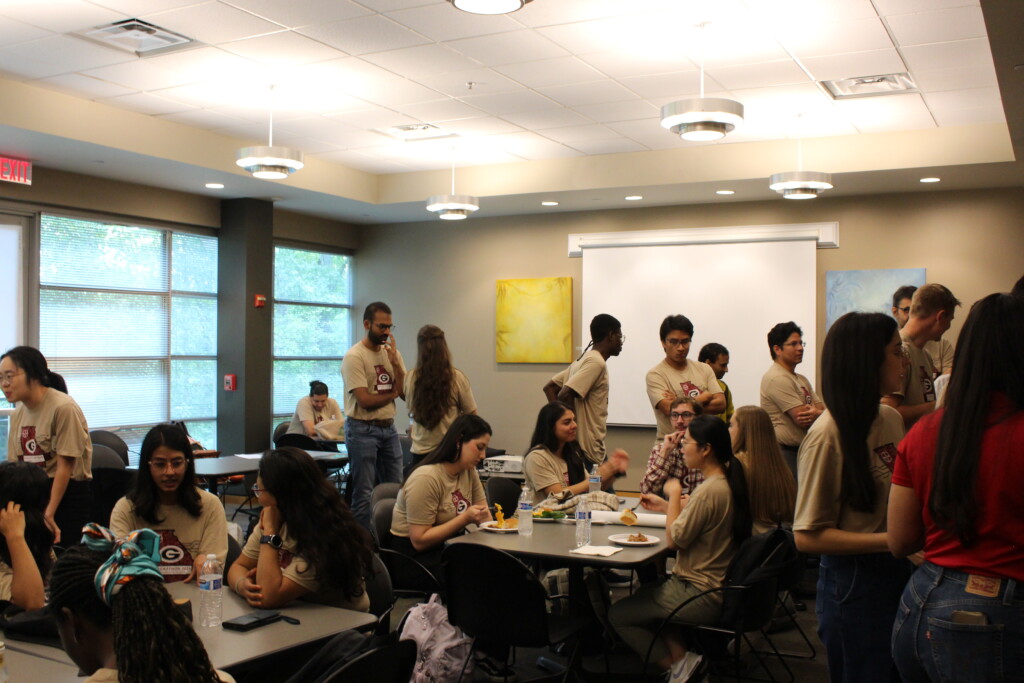 students sit at tables and eat or talk, most in matching tan shirts, while others stand in line at the back of the room to get food.