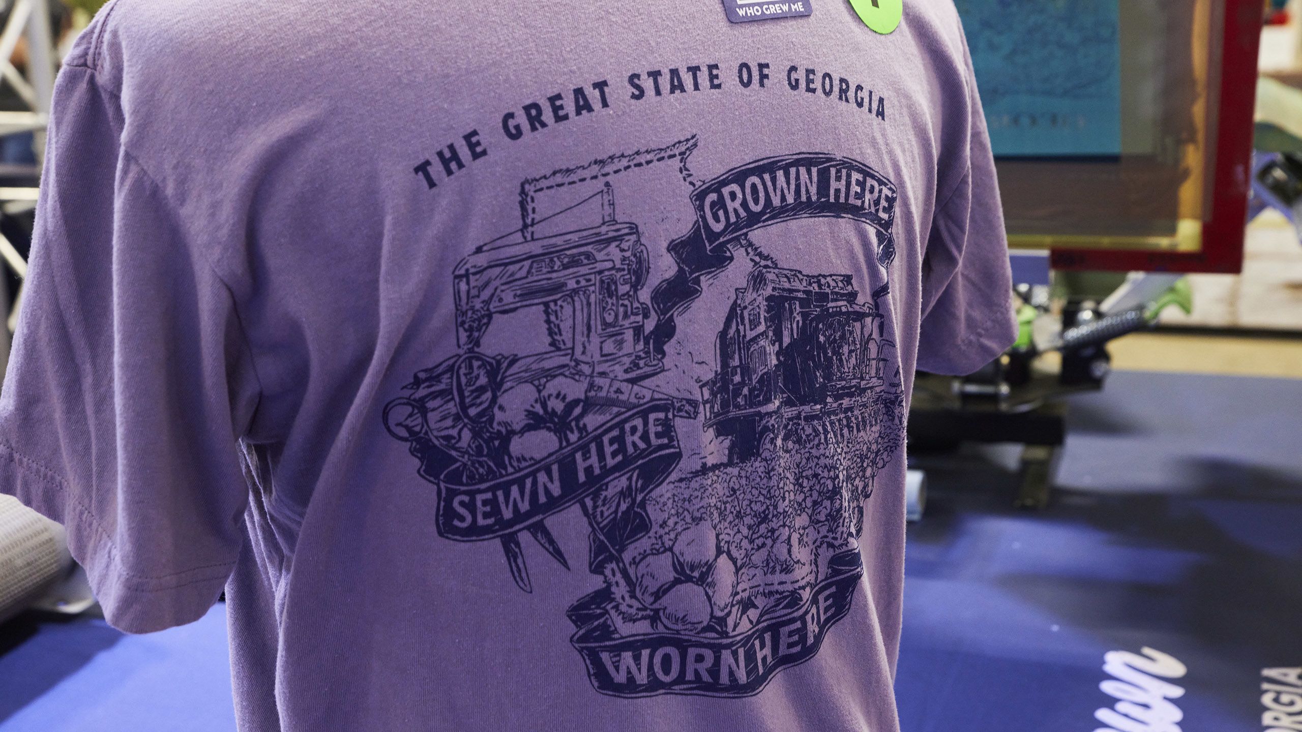 Purple t-shirt with "The Great State of Georgia" and illustrations of cotton, a sewing machine, and text "Grown Here, Sewn Here."