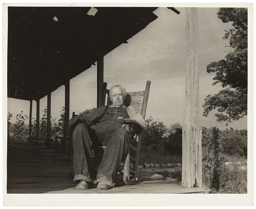 An elderly man sits in a rocking chair on a wooden porch, wearing overalls and a shirt, with trees and an open field in the background.