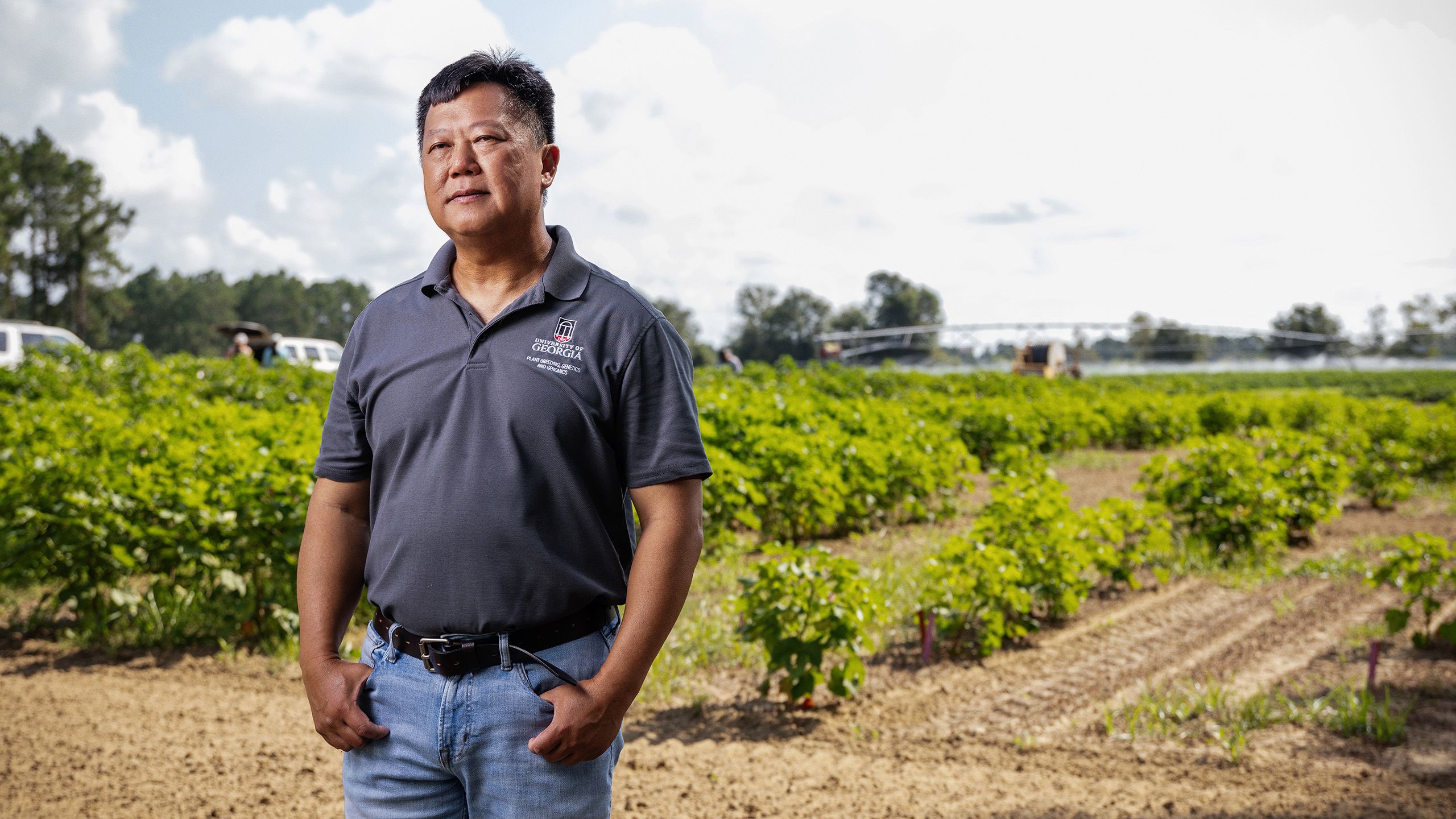 Man in a dark polo shirt stands in a cotton field, hands in pockets, with green plants and a partly cloudy sky behind him.