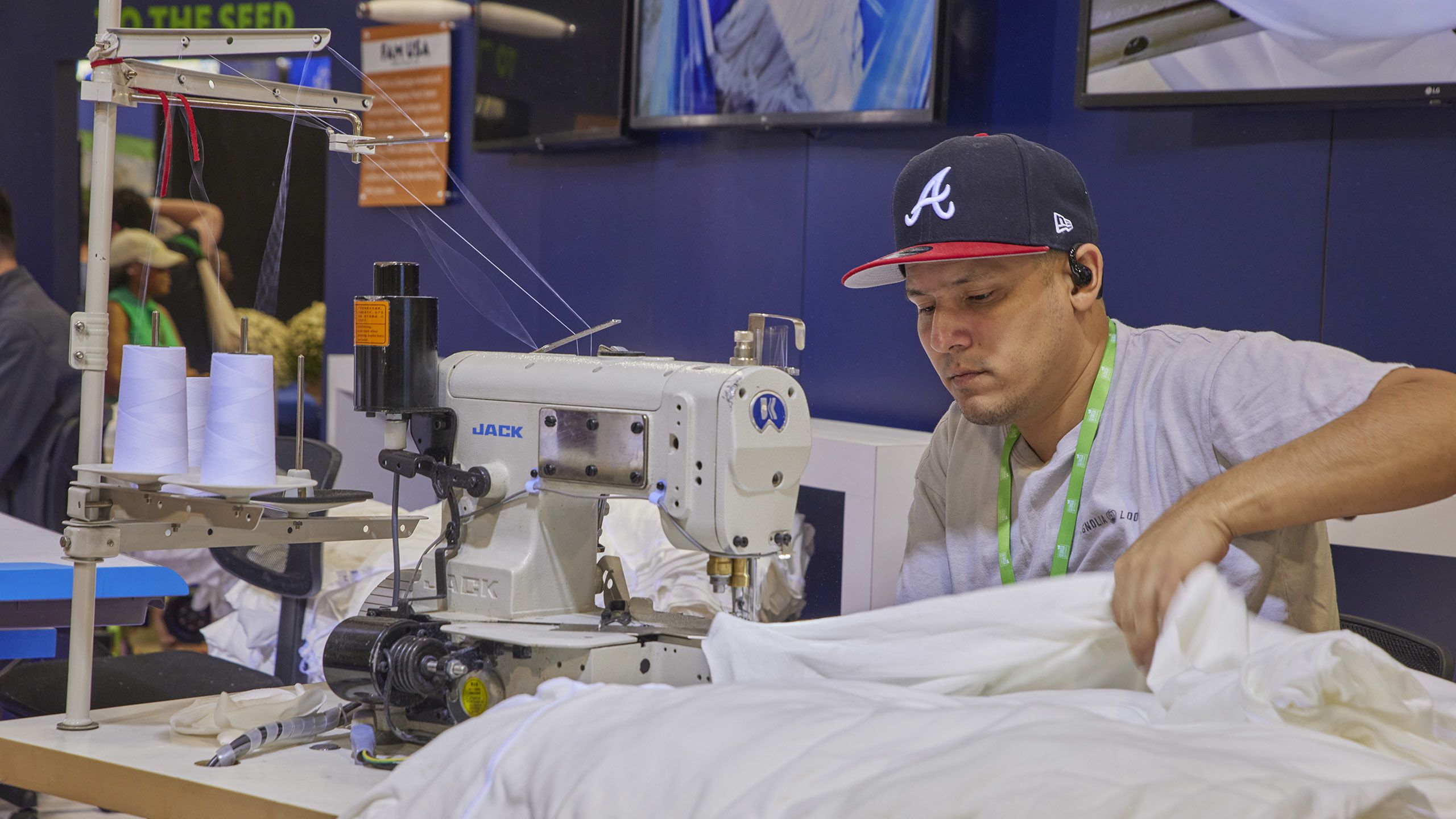 Man in a baseball cap operates a sewing machine, stitching white fabric in a workshop setting.