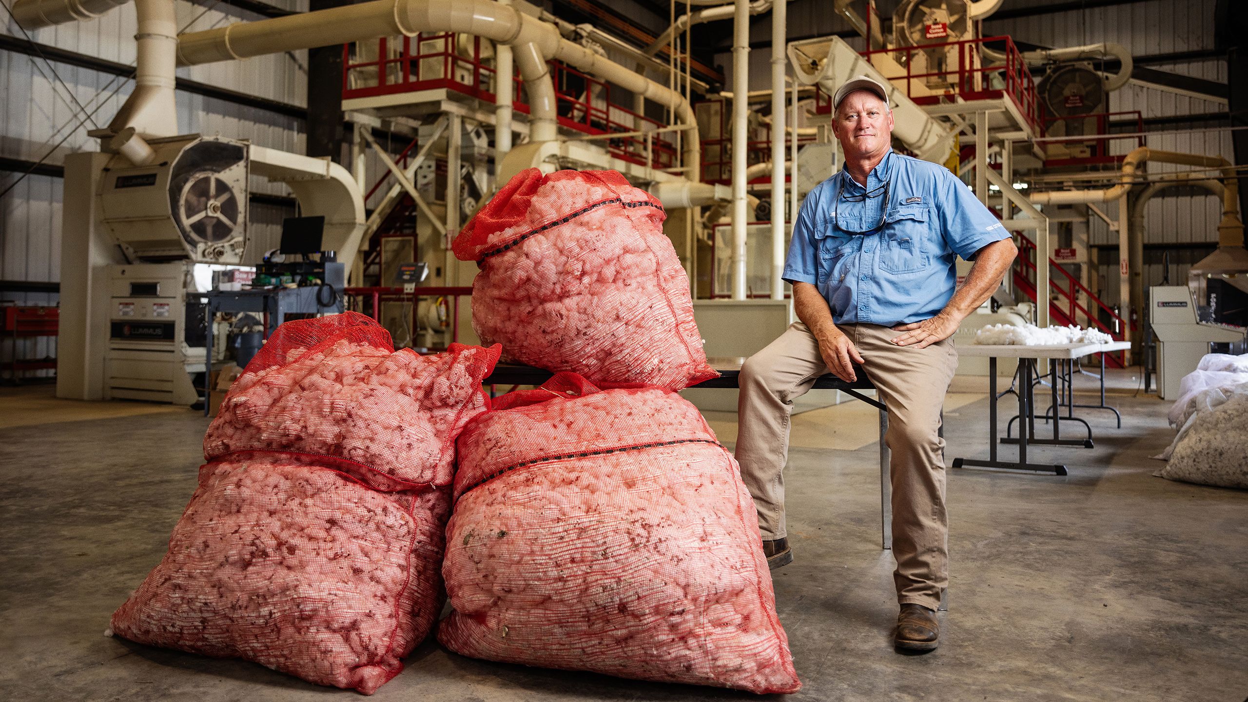 Man in a blue shirt sits on a bench beside large mesh bags of cotton in a gin facility with industrial machinery.