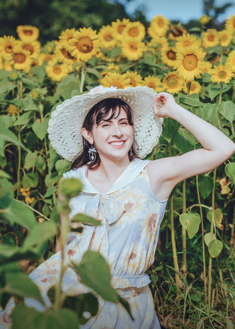 A smiling young woman stands in a field of sunflowers, wearing a dress with a sunflower print. She has a white sunhat on, and one arm is holding the brim.
