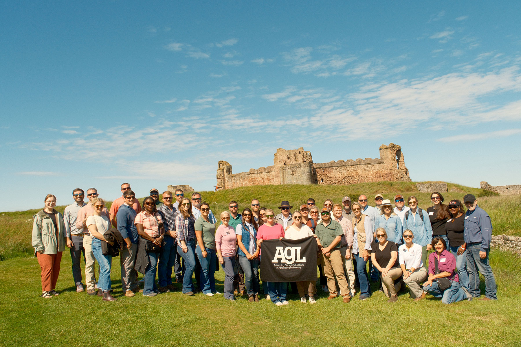A large group of people standing together outdoors on a grassy hillside with a historic stone castle ruin in the background under a bright blue sky with scattered clouds. The group is posing for a photo, and two people in the front center are holding a black banner with white text that reads “AGL.”