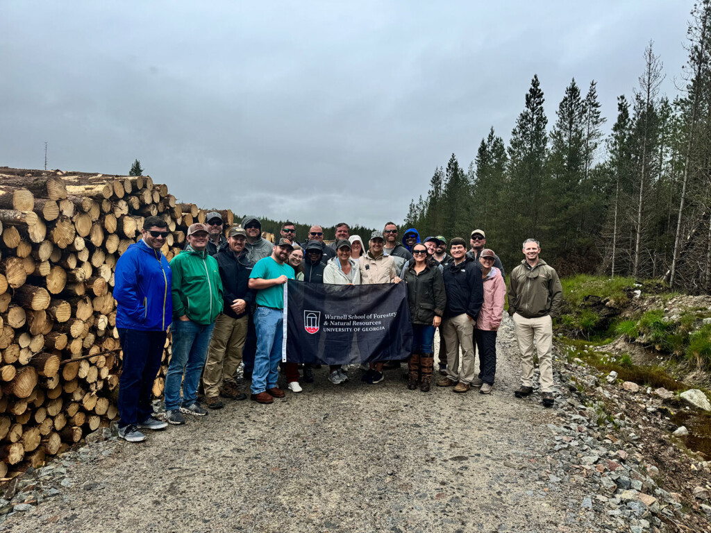 Group of people standing on a dirt road in a forested area holding a Warnell School of Forestry banner.