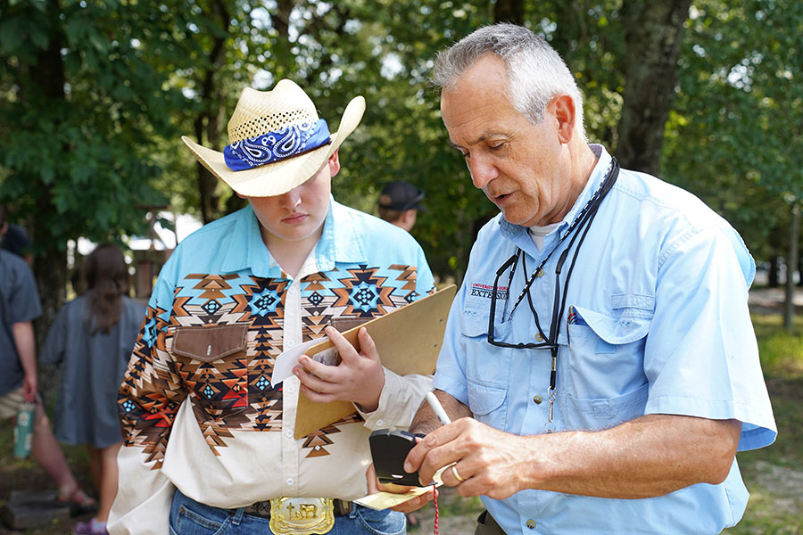A young man in a cowboy hat and Western patterned shirt listens attentively to a man in a blue shirt with the UGA Cooperative Extension logo above his right shirt pocket who is consulting a measuring device. (Photo by Josie Smith)