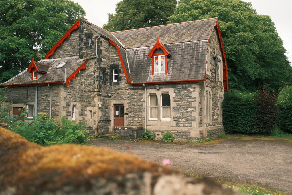 Historic stone cottage with steep roof and red trim surrounded by trees.