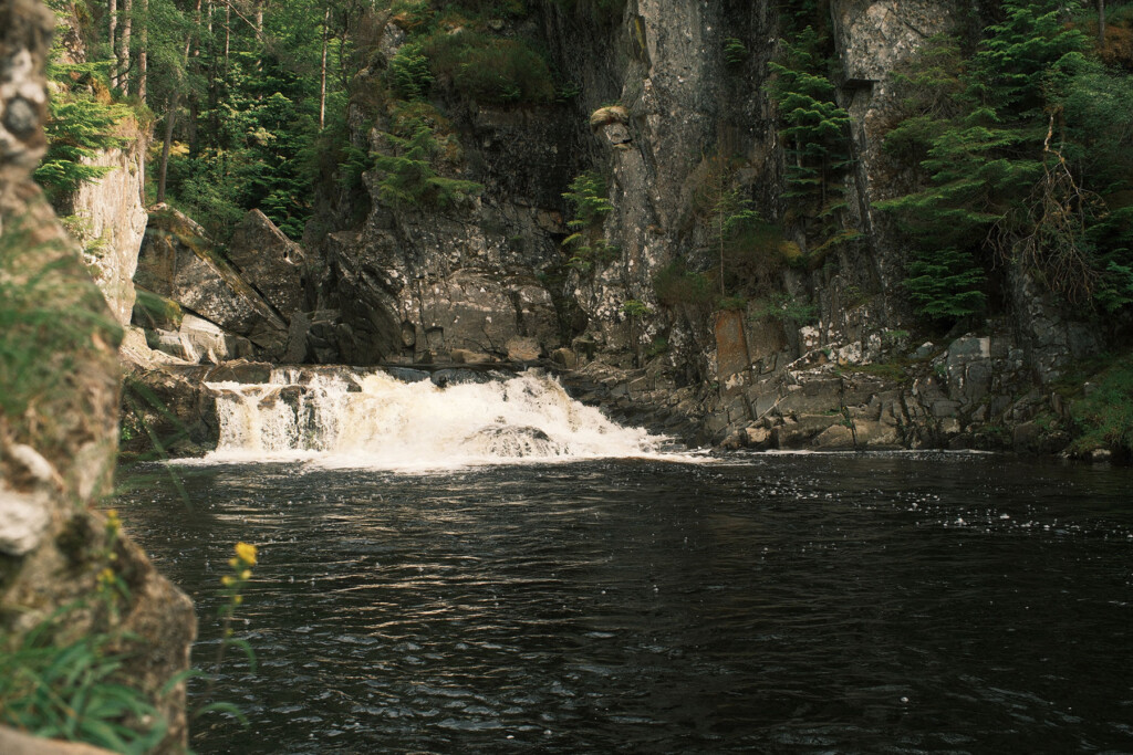 Small waterfall flowing into a dark pool surrounded by rocky cliffs and evergreen trees.