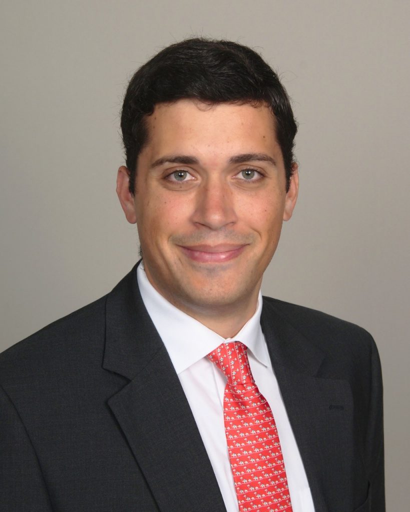 Ford Ramsey has short dark hair and wears a dark suit jacket, white dress shirt and a red patterned tie smiles at the camera against a neutral gray background.