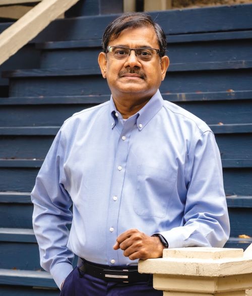 Gopi Munisamy wears glasses and a light blue button-down shirt stands outdoors in front of dark blue wooden steps, resting one hand on a light-colored railing and looks toward the camera.