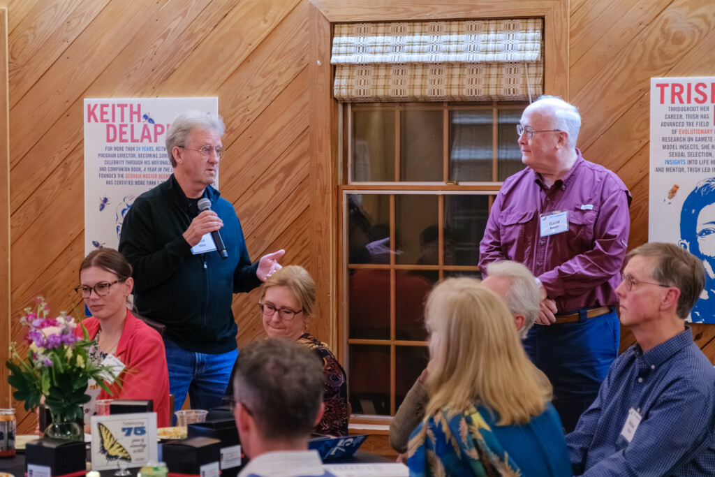 A person holds a mic and addresses another standing person as well as a room full of seated people.