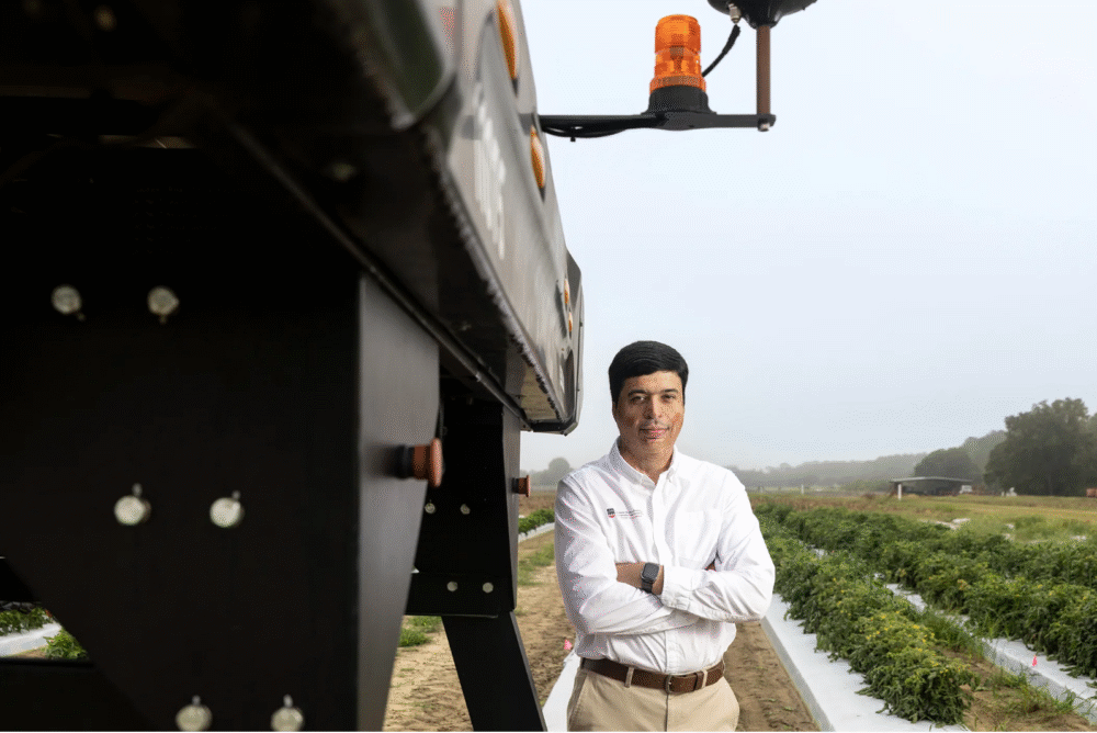 A man wearing a white shirt and khaki pants stands with his arms crossed next to a large piece of farm equipment in a field setting.