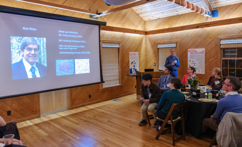 A person holding a microphone addresses the room alongside a projected slide that reads 'Ken Ross' and details where he received his various degrees.