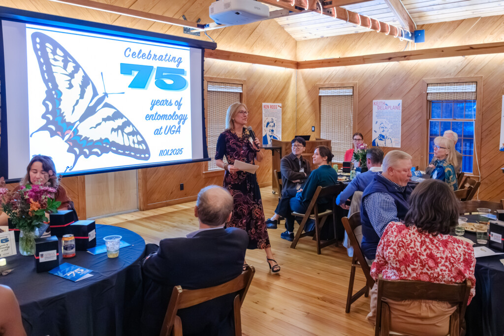 A speaker stands in front of a projected presentation and addresses a seated audience through a mic. The test on the presentation slide reads "Celebrating 75 years of entomology at UGA." and the date of the event, November 1st,  2025.