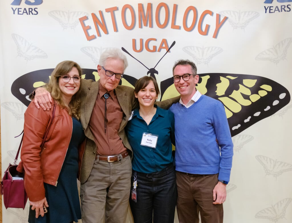Four people pose for a photo in front of a backdrop of giant butterfly wings. Text on the backdrop reads 'Entomology UGA'.