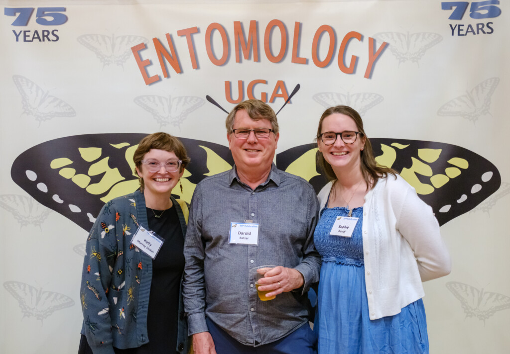 Three people pose for a photo in front of a backdrop of giant butterfly wings. Text on the backdrop reads '75 years' and 'Entomology UGA'.