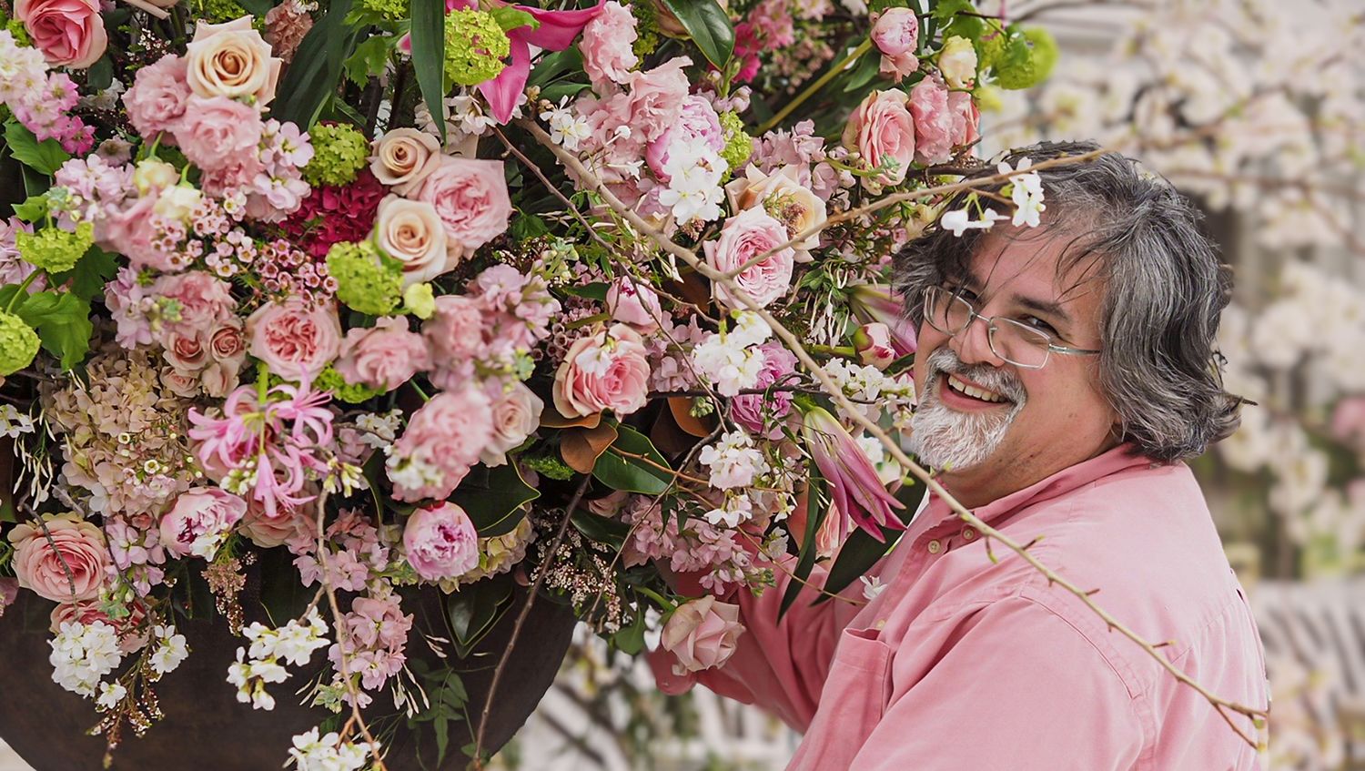 Man in a pink shirt holding a large, vibrant floral arrangement with pink, white, and green flowers, surrounded by blossoms.