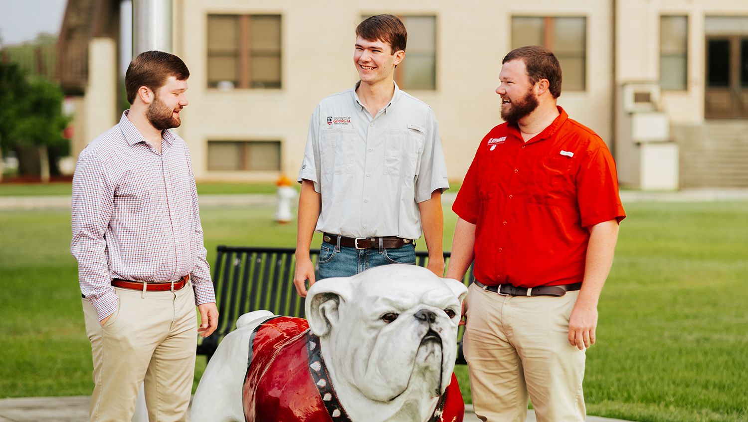 Three men standing outdoors on a campus lawn, smiling near a white bulldog statue wearing a red jacket.
