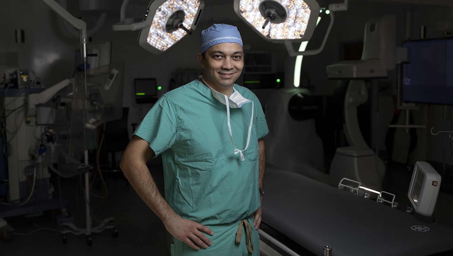 Surgeon in green scrubs and blue cap standing in an operating room, with surgical lights and medical equipment in the background.