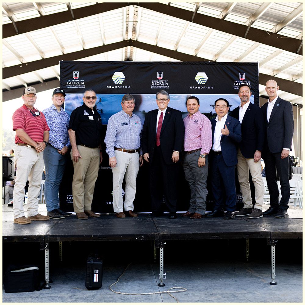 Group of eight men standing together at an outdoor event, dressed in business casual attire, with UGA banners in the background.