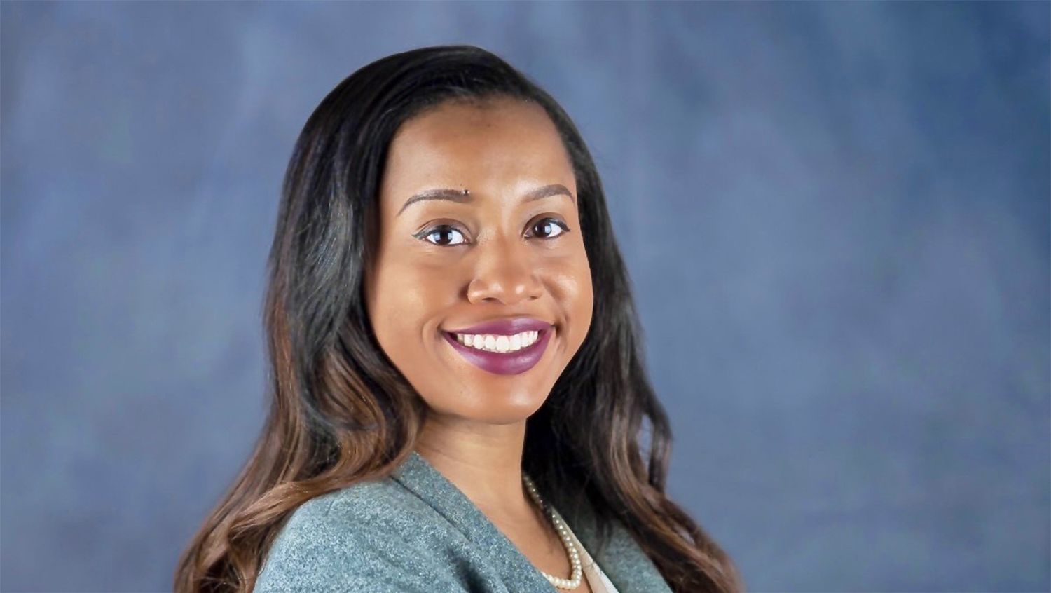 Woman with long dark hair smiling, wearing a gray top and pearl necklace.