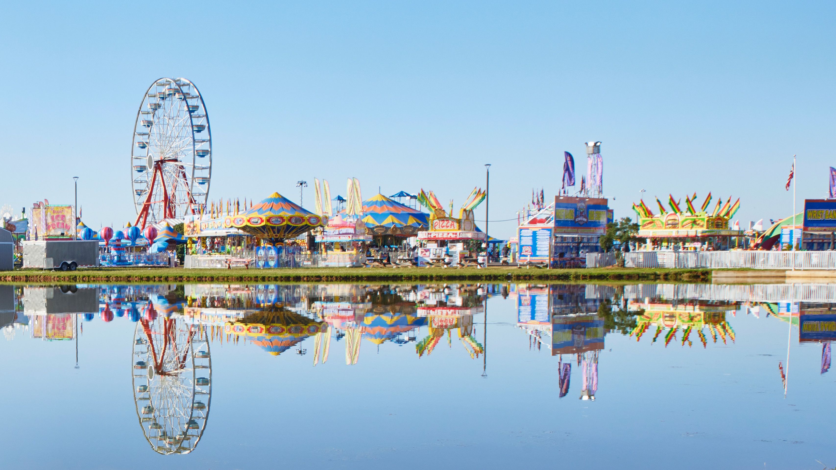 Colorful fairground with a Ferris wheel, rides, and food booths reflected in calm water under a clear blue sky.