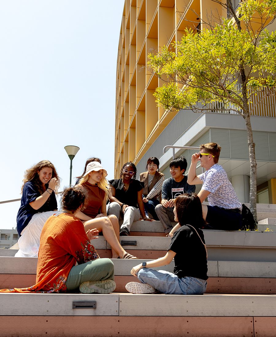 Group of students sitting on outdoor steps, engaged in conversation, with a modern yellow building and green tree in the background.