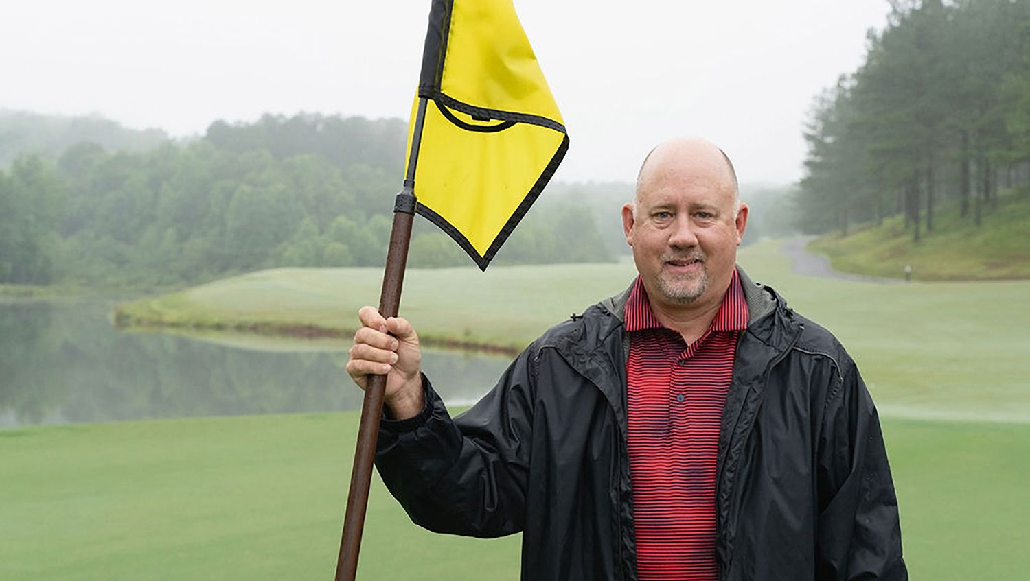 Man holding a yellow golf flag on a green course, wearing a black jacket and red striped shirt, with a misty background.