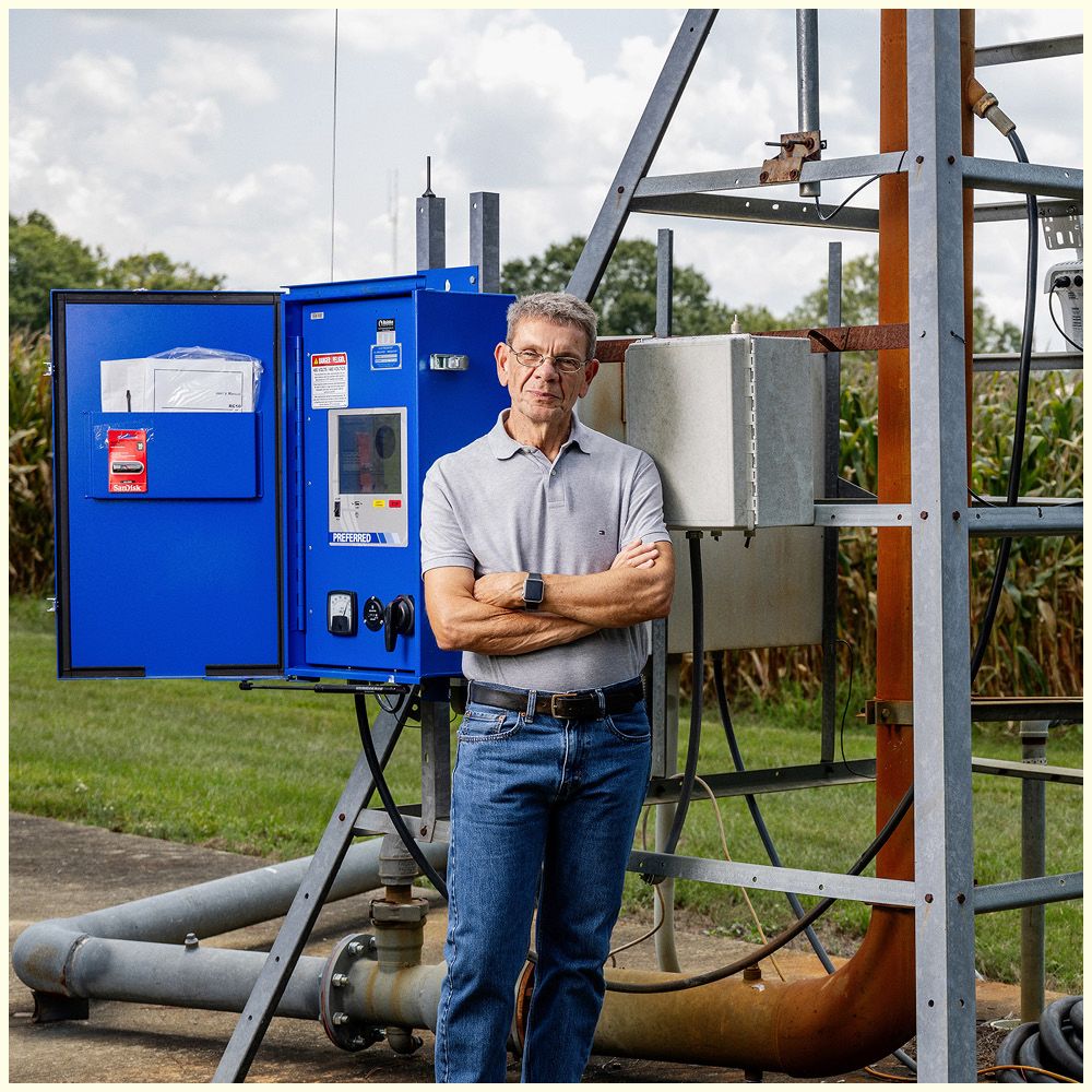 Man standing with arms crossed in front of a blue agricultural machine, wearing a light shirt and jeans, outdoors.