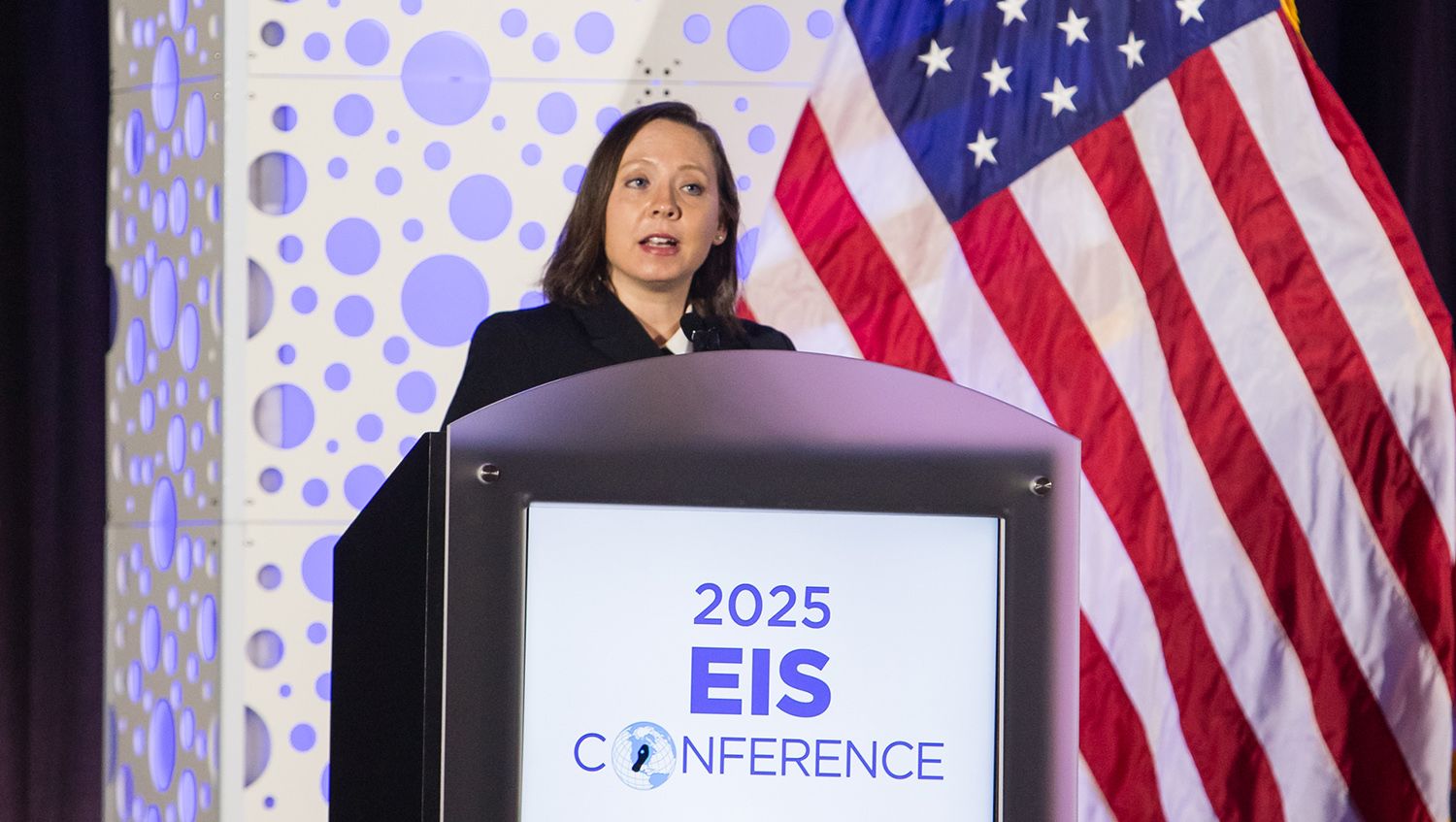 Woman speaking at a podium labeled "2025 EIS Conference," with an American flag and a patterned backdrop behind her.