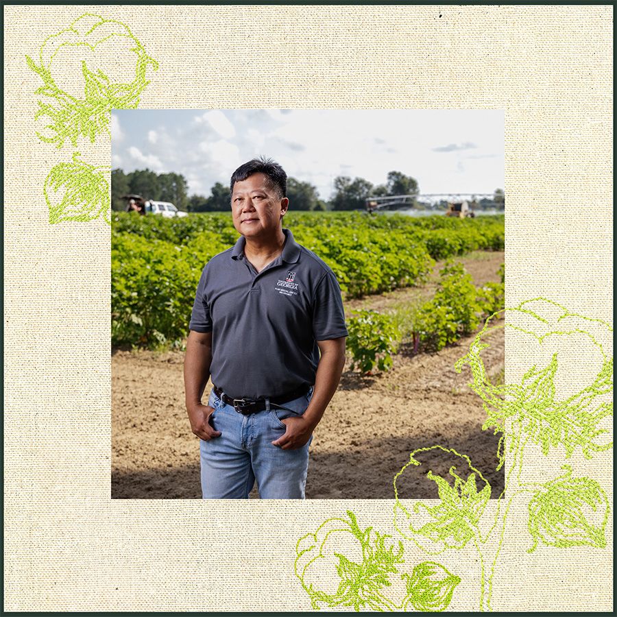 Man standing in a field of green crops, wearing a dark polo shirt and jeans, with farm equipment visible in the background.