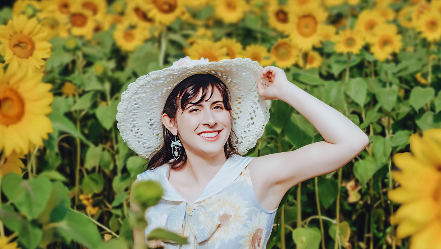 Woman in a white hat and floral dress smiling in a sunflower field, adjusting her hat with one hand.