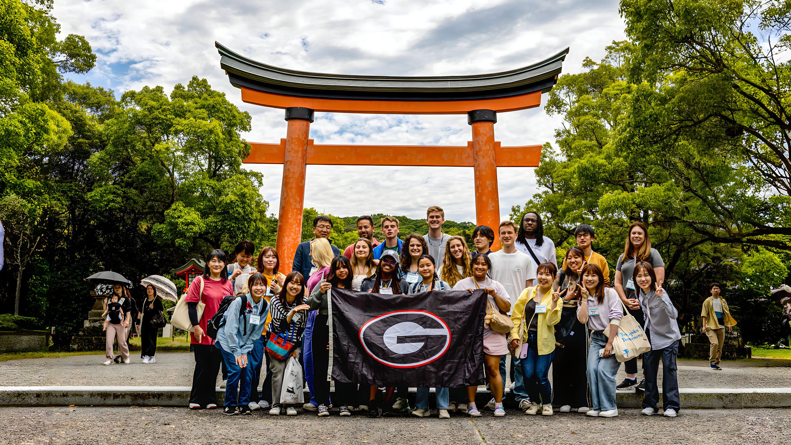 Group of UGA students posing in front of a large orange torii gate at a Shinto shrine, holding a UGA banner, surrounded by green trees.