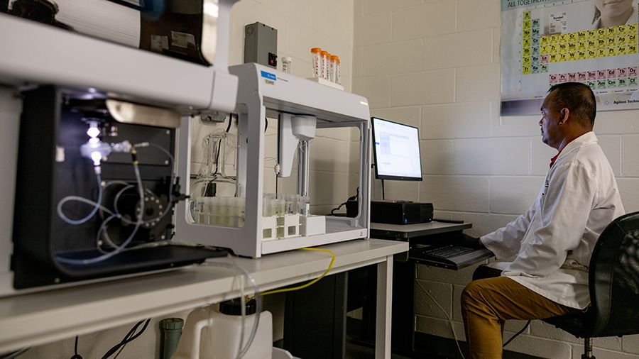 A scientist in a lab coat sits at a computer, monitoring a machine analyzing soil samples in a laboratory.