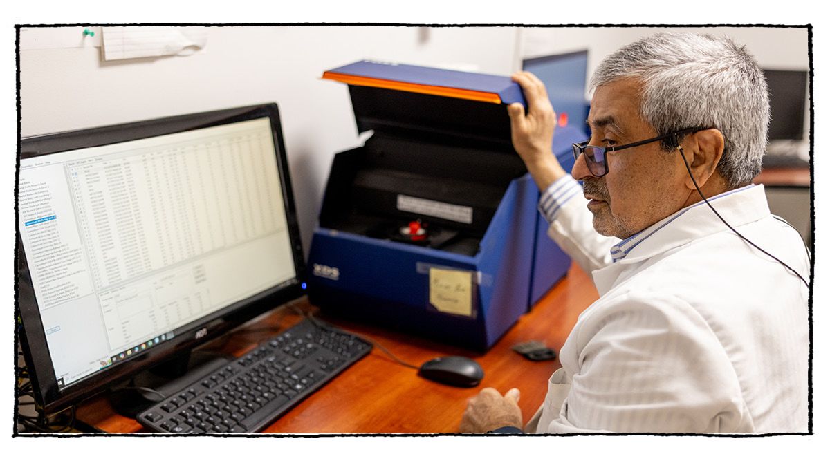 A man in a lab coat gazes at a computer monitor as he closes a tabletop wet chemistry device