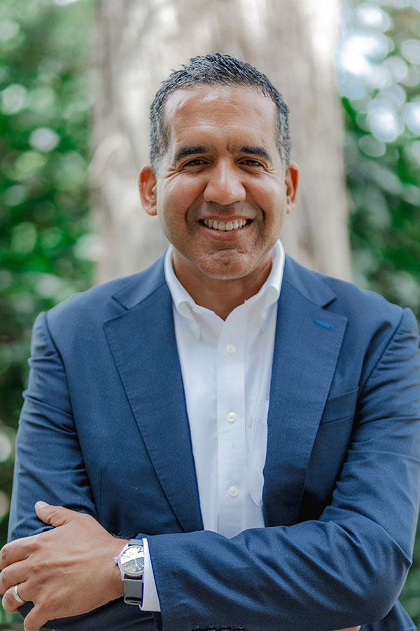 A smiling man with dark hair wearing a blue blazer and light blue dress shirt stand with his arms crossed casually in front of him.