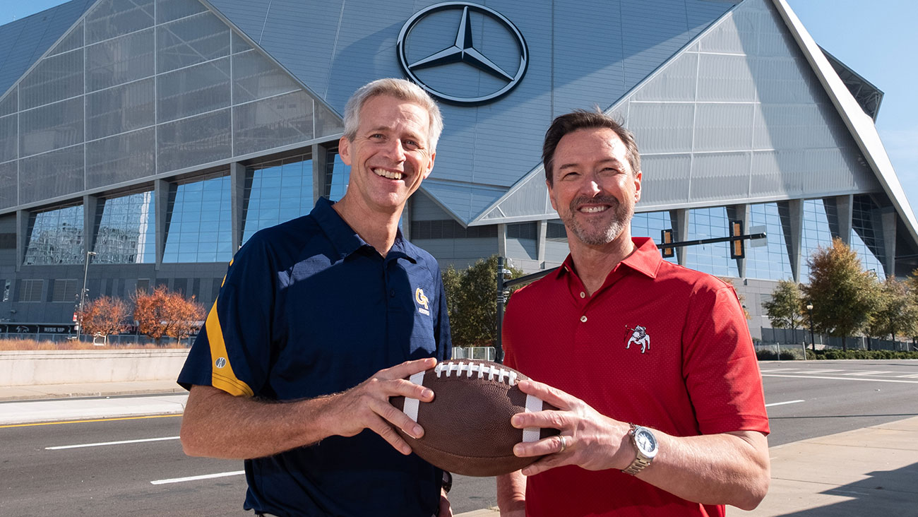 Two men stand smiling in front of Mercedes-Benz Stadium, one wearing a navy Georgia Tech shirt and the other wearing a red University of Georgia shirt. They hold a football together as they pose for the photo on a sunny day.
