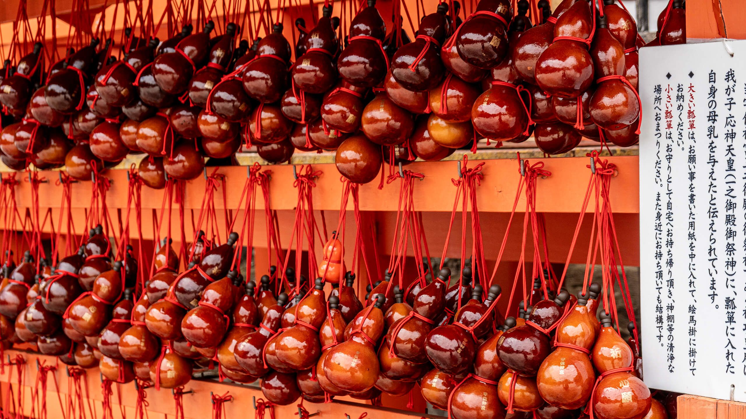 Rows of red lacquered gourd-shaped charms hanging on a wooden rack, with a sign in Japanese explaining their use for wishes at a shrine.