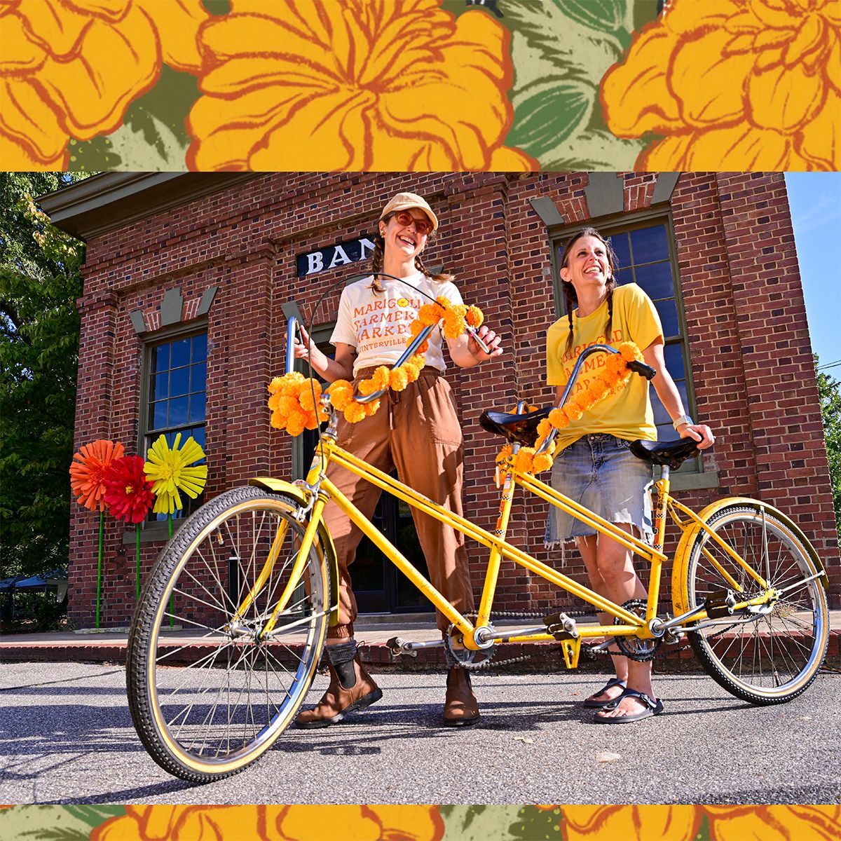 Two women smiling beside a yellow tandem bicycle decorated with marigold flowers, standing in front of a brick building.