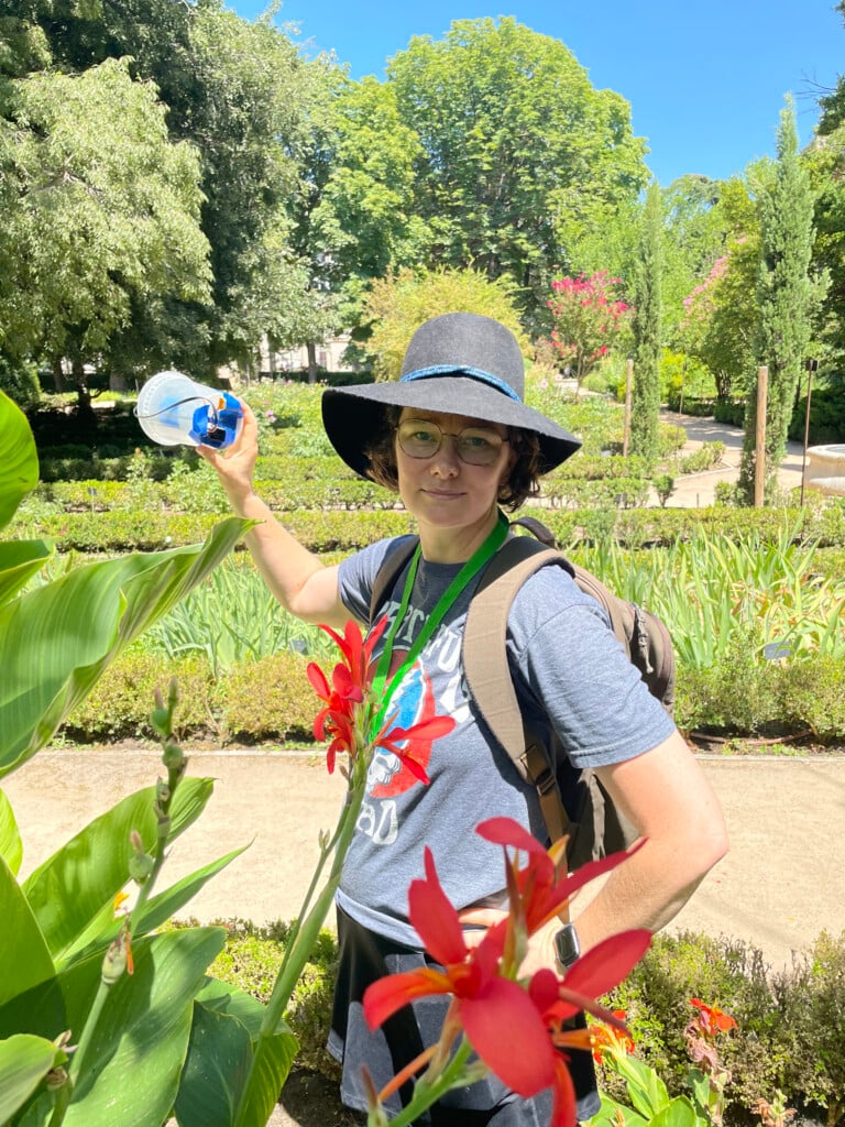 Salzman wears a hat and backpack and holds a device to measure carbon dioxide while standing in a garden with green plants, trees and red blooming flowers in the foreground.