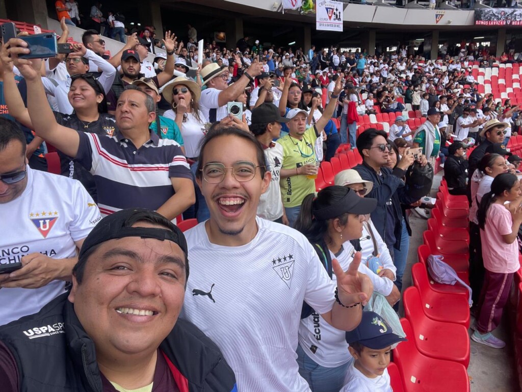 Two people in the stands of a sporting event smile at the camera with crowds around them.