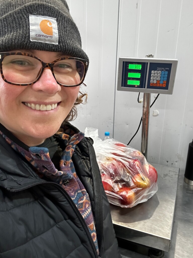A close up selfie of a woman standing in front of a bag of apples on a scale.
