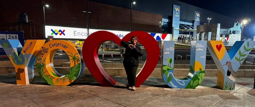 A photo of a woman holding her hands in the shape of a heart in front of a sign that reads YO heart UY.