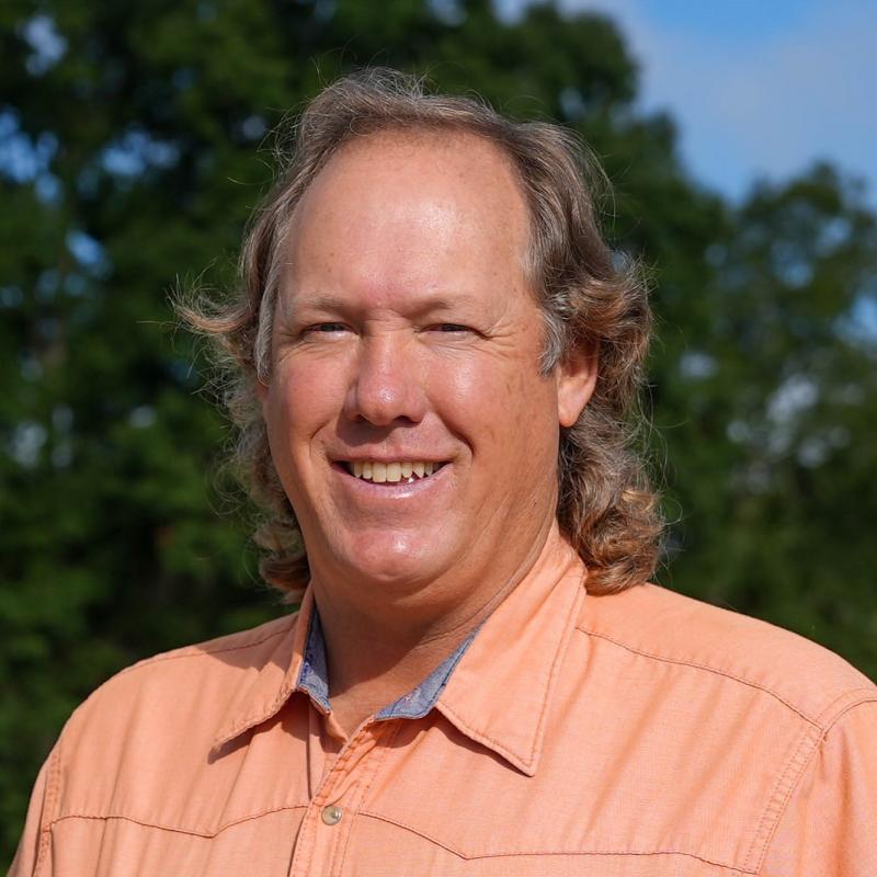 Wearing a light orange collared shirt, John Gassett smiles while standing outdoors