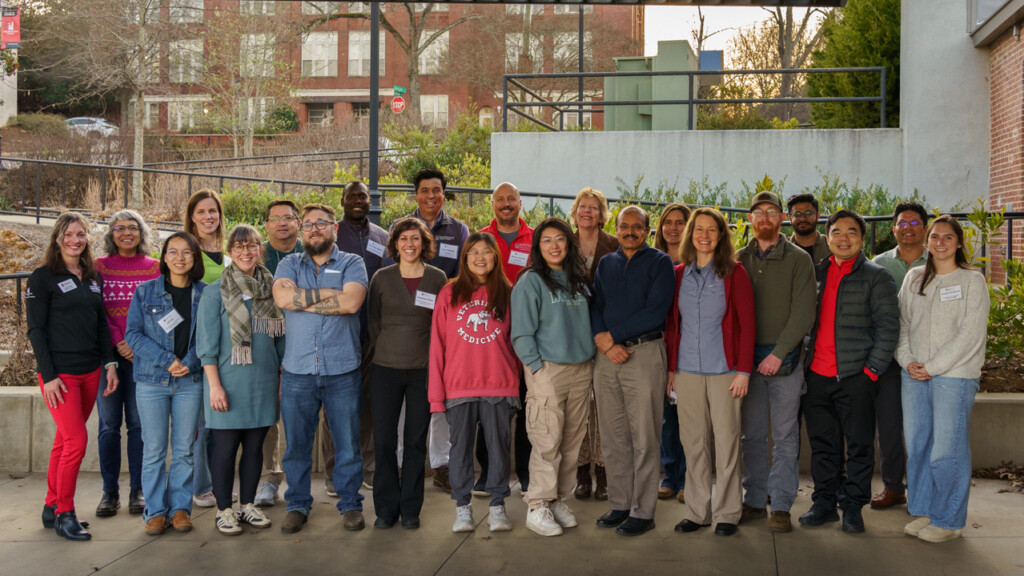 A large group of people standing outdoors smiling with shrubbery and brick buildings in the background.