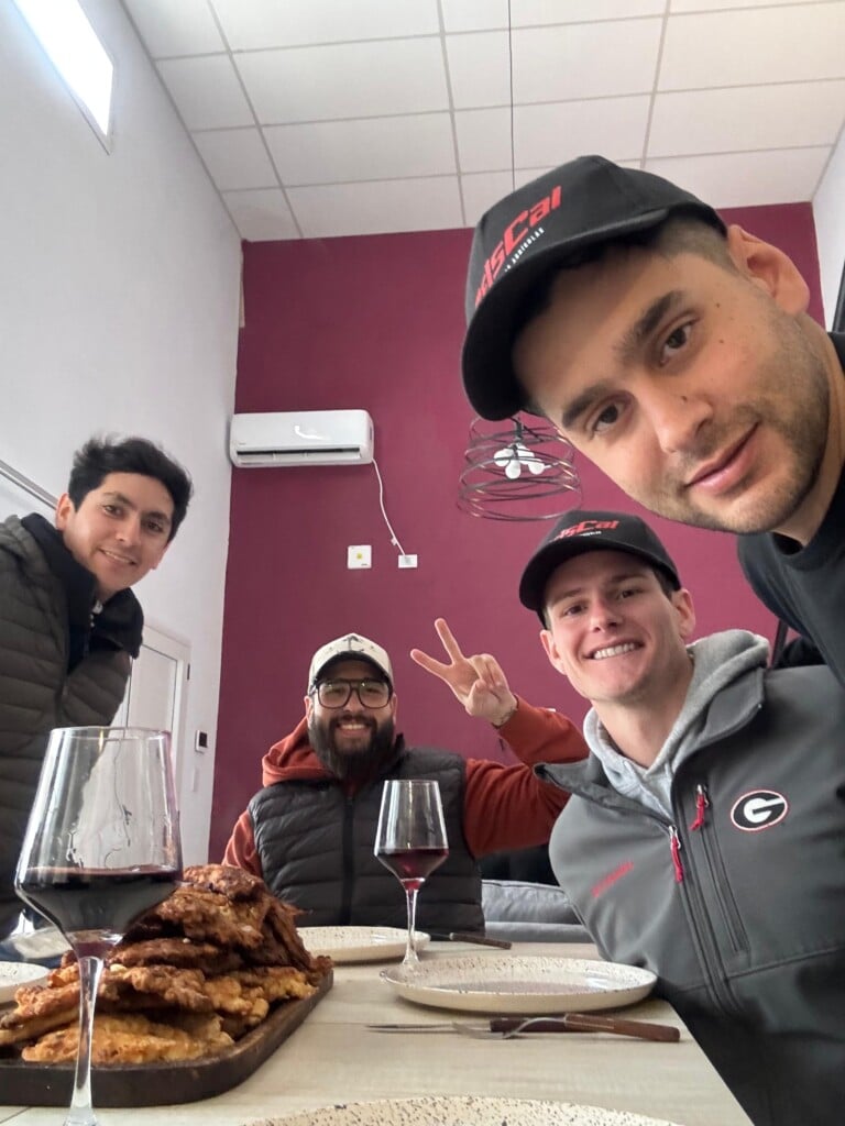 Four people at a dinner table smile at the camera. One holds up a peace sign with two fingers.