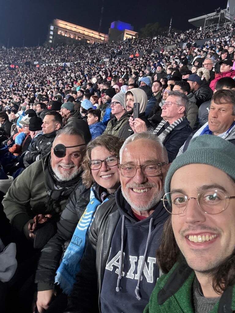 Four people in the foreground smile at the camera with a crowd behind them watching a soccer game.