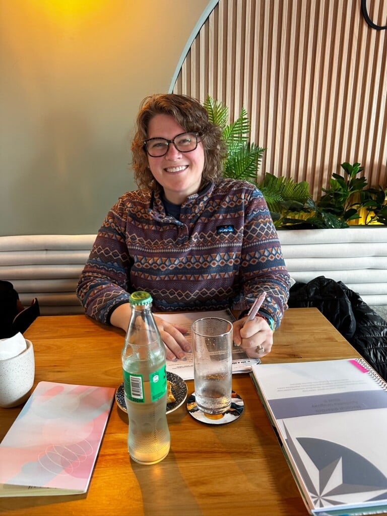 A woman in a multicolored sweater smiles at the camera while sitting in a cafe.
