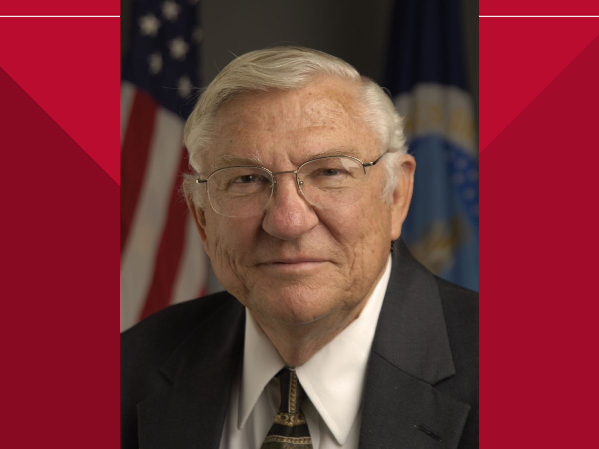 Portrait of Dr. Gale Buchanan wearing a dark suit, white shirt, and patterned tie. He is in front of two flags, including the American flag, with a neutral background.